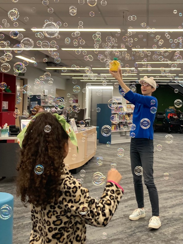 In front of the children's desk in a library, Ingrid Christina holds up a pineapple bubble machine that is filling the room with bubbles. They are wearing a white hat, blue shirt, and black pants. In the foreground, a child is viewed from behind catching bubbles.